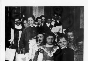 Schoolchildren of different ages are lined up, holding documents in their hands. Original caption reads "Well-Doers Waiting for the Principal's Commendation"