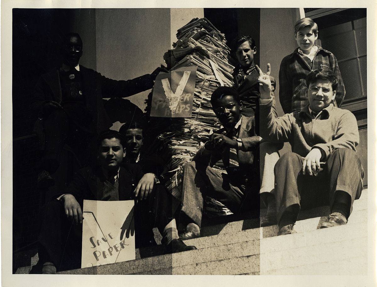A group of Black and white children sit on steps beside a large stack of paper, making &quot;V&quot; signs with their hands and holding campaign signs