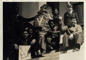A group of Black and white children sit on steps beside a large stack of paper, making &quot;V&quot; signs with their hands and holding campaign signs