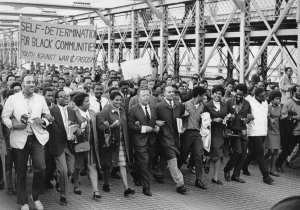 Black education advocates march over the Brooklyn Bridge together, arms linked. A large sign reads "Self-Determination for Black Communities, Youth Against War & Fascism"