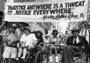 Wheelchair users and a little person march in a parade with a banner