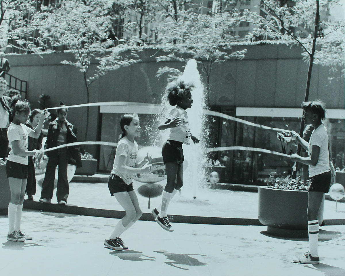 Girls jump rope near Lincoln Plaza. Two girls turn the ropes and the others jump. One girl is mid-air and a fountain behind her sprays upward. The other girl has knees bent, ready to jump.