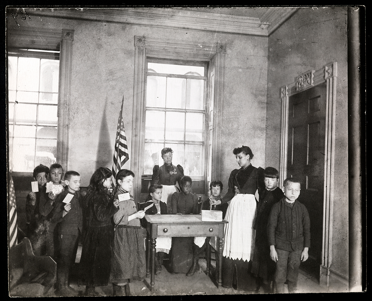 Around 10 schoolchildren of various races and genders line up with ballots in hand. One of two women teachers is placing a ballot in a box on a desk.