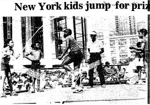 Black children jump rope in front of a crowd of seated people. A referree watches closely.