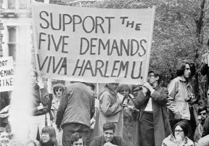 College students hold signs at an outdoor rally. A large banner reads, &quot;Support the Five Demands - Viva Harlem U.&quot;