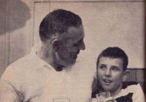 A white middle-aged man sits next to his son. They hold drinks and look at each other. The father has his arm around his son, and both are smiling.